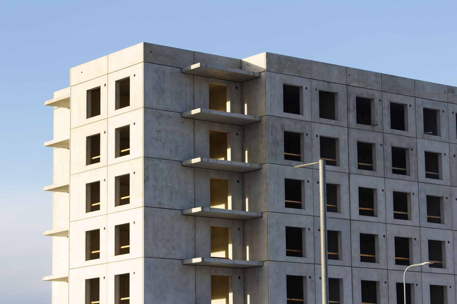 Sunlit concrete building under construction with unfinished window openings and projecting balconies against a clear blue sky.